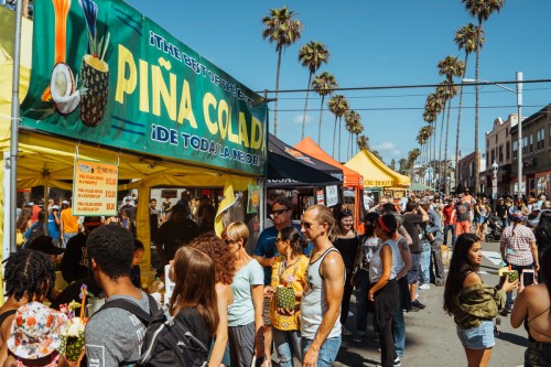 A bustling street market with colorful stalls, palm trees, and crowds enjoying food and drinks under a clear blue sky.