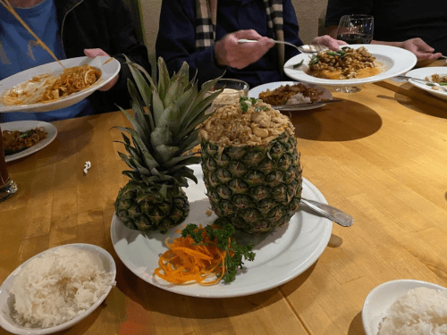 A pineapple filled with fried rice, garnished with herbs, surrounded by plates of food and rice on a wooden table.