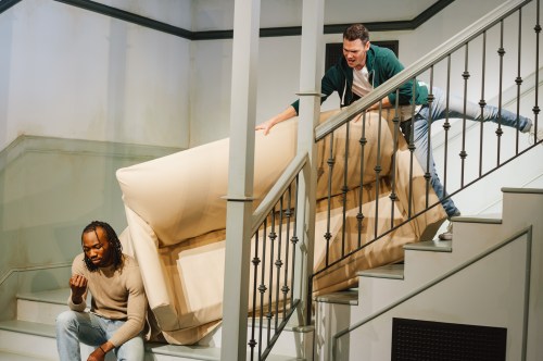 Two men are moving a large couch up a staircase, one struggling while the other assists from above.