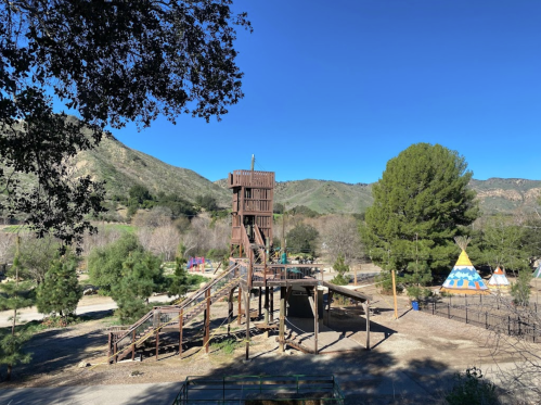 A wooden play structure surrounded by trees and mountains under a clear blue sky. Tents are visible in the background.