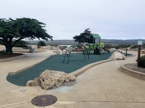 A playground with green equipment, surrounded by rocks and trees, near a calm water view on a cloudy day.