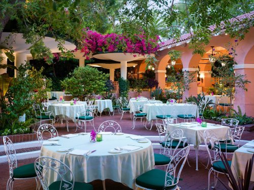 A charming outdoor restaurant with white tables, green chairs, and vibrant flowers in a lush garden setting.