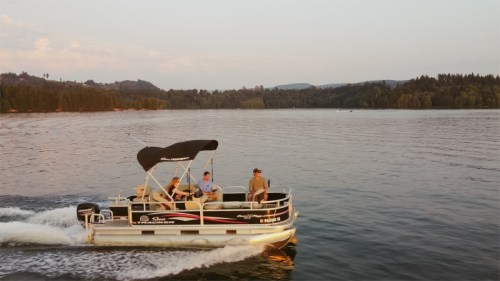 A pontoon boat with four people cruising on a calm lake during sunset, surrounded by trees and hills.