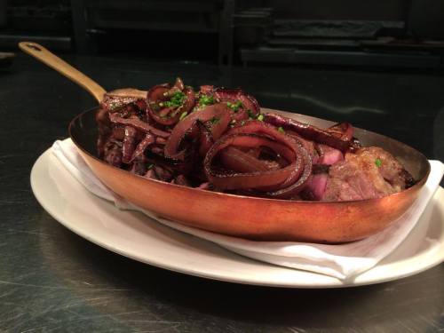 A copper serving dish filled with sliced steak topped with caramelized onions and garnished with parsley.
