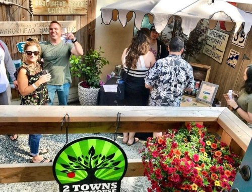 A lively outdoor gathering with people socializing, a vendor booth, and colorful flowers in a rustic setting.