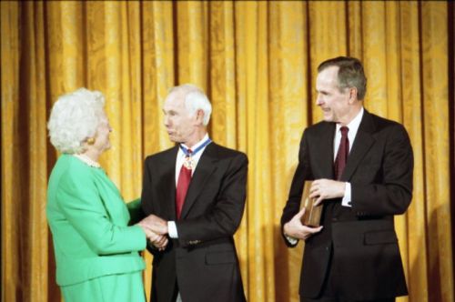 A man receives an award from a woman and a man in formal attire, with a golden curtain in the background.