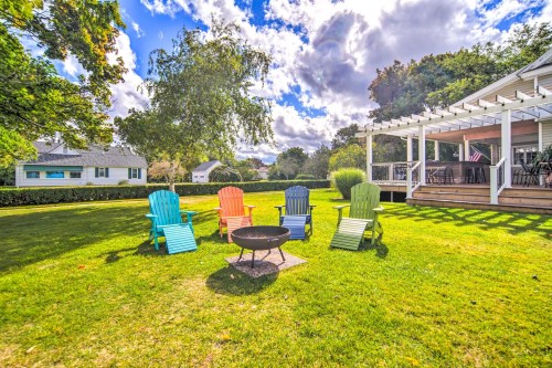 Colorful Adirondack chairs surround a fire pit on a grassy lawn, with a house and trees in the background under a cloudy sky.
