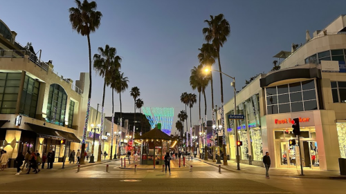 A lively street scene at dusk, lined with palm trees, shops, and colorful lights in the background.