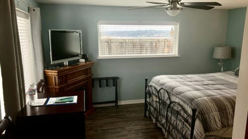 Cozy bedroom with a bed, TV on a stand, and a window with blinds, featuring light blue walls and wooden flooring.
