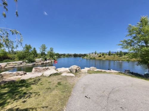 A serene park scene featuring a calm lake, rocky shoreline, and lush greenery under a clear blue sky.
