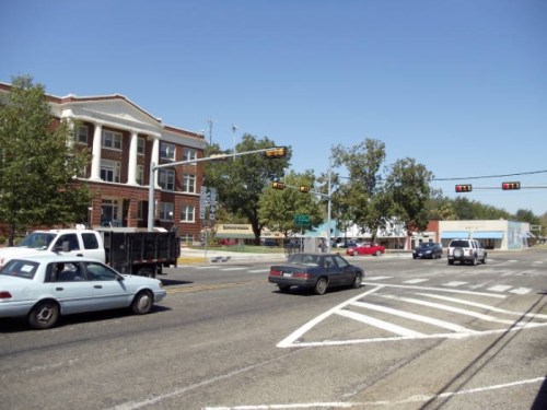 A busy intersection with traffic lights, featuring a brick building and parked cars under a clear blue sky.