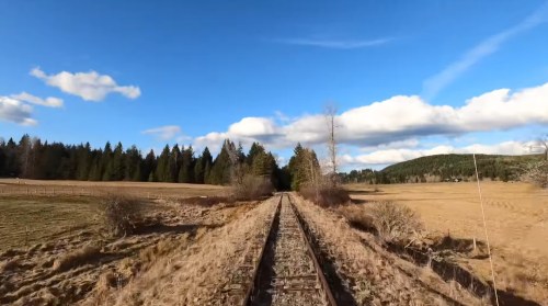 A view of a railway track stretching through a grassy field, surrounded by trees and under a blue sky with clouds.