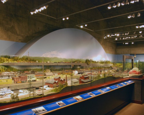 A detailed model of a town displayed in a museum, featuring buildings, water, and a scenic backdrop.