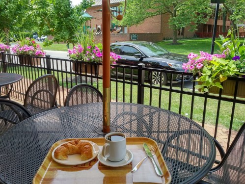 A table set with a croissant and coffee, surrounded by flowers and greenery, with a car parked in the background.