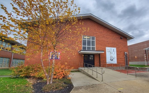 A brick building with a ramp, surrounded by autumn trees and cloudy skies. A mural is visible on the wall.