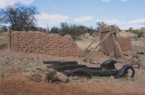 Ruins of an adobe structure with a wooden support, surrounded by dry grass and scattered debris.