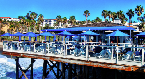A seaside restaurant on a pier with blue umbrellas, surrounded by palm trees and ocean waves.