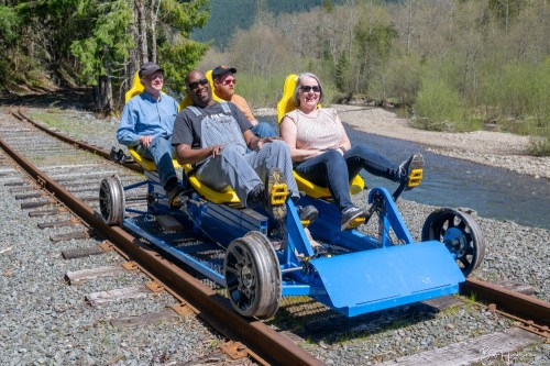 Four people sit on a blue rail bike on a gravel track, surrounded by trees and a river in a scenic outdoor setting.