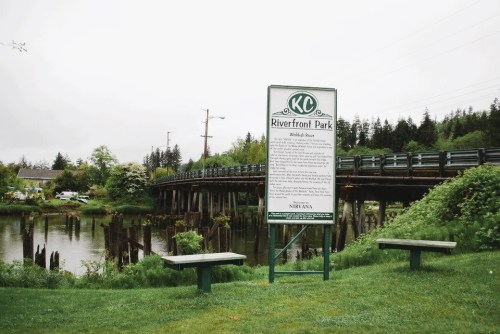 Sign for KC Riverfront Park near a bridge, with benches and greenery by the water on a cloudy day.