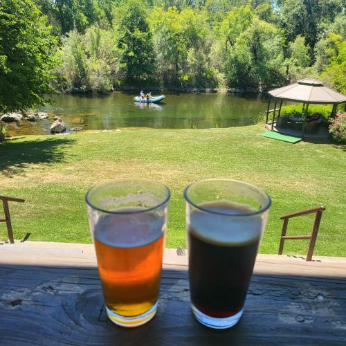 Two glasses of beer sit on a wooden table, overlooking a serene river and people in a boat amidst lush greenery.