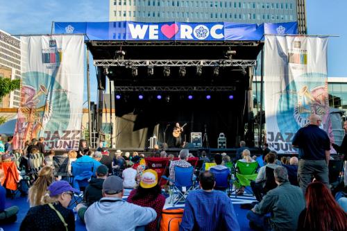 A musician performs on stage at an outdoor festival, with an audience seated on the ground in front. Banners display "WE ♥ ROC."