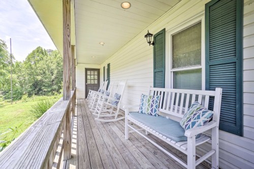 A porch with white rocking chairs and benches, featuring decorative pillows, overlooking a green landscape.