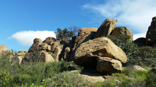 Rocky landscape with large boulders and green vegetation under a blue sky with scattered clouds.