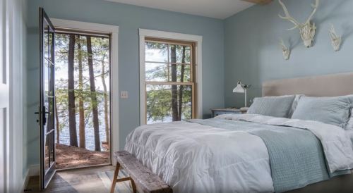 Cozy bedroom with a light blue wall, a bed, and a door opening to a scenic view of trees and water.