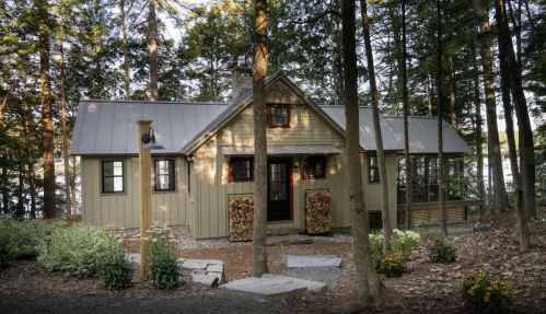 A cozy cabin nestled among trees, featuring a metal roof, stone chimney, and stacked firewood by the entrance.