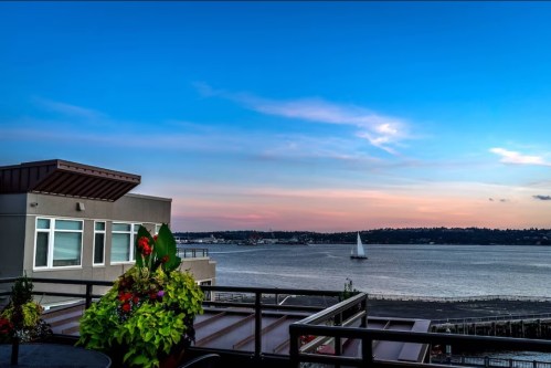 A scenic view of a waterfront with a sailboat, colorful flowers in the foreground, and a vibrant sky at sunset.