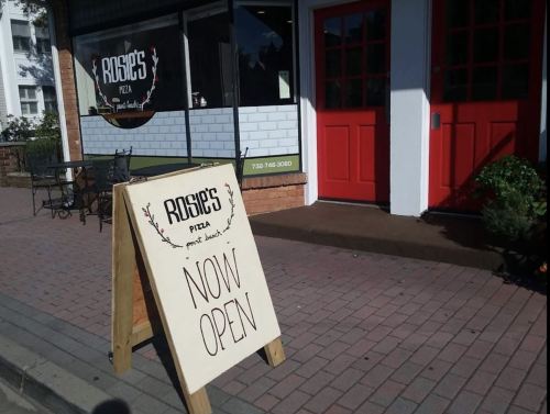 A sidewalk sign outside Rosie's Pizza reads "NOW OPEN," with the restaurant's entrance visible in the background.