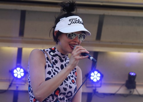A singer in a leopard print outfit and visor performs on stage, holding a microphone with blue lights in the background.