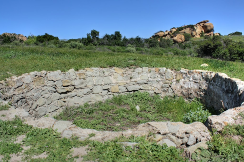 A circular stone structure surrounded by green grass and rocky hills under a clear blue sky.