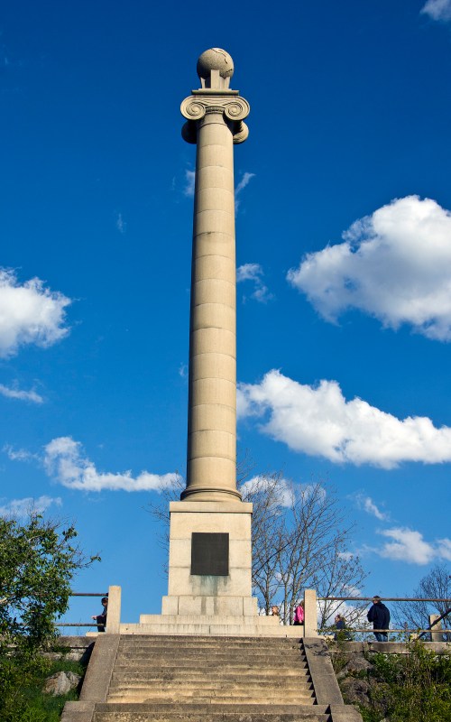 A tall stone monument with a spherical top, surrounded by trees and a blue sky with clouds. People are walking nearby.