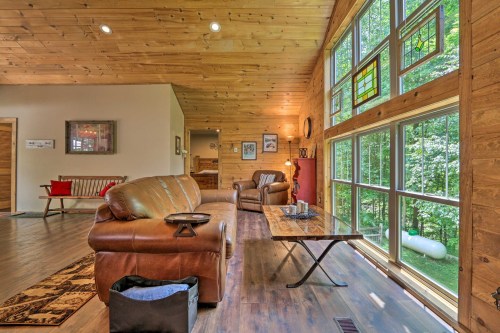 Cozy living room with wooden walls, large windows, a brown sofa, and a coffee table, surrounded by greenery outside.