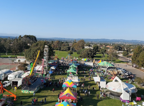 Aerial view of a vibrant fair with colorful tents, rides, and crowds in a park setting under a clear blue sky.