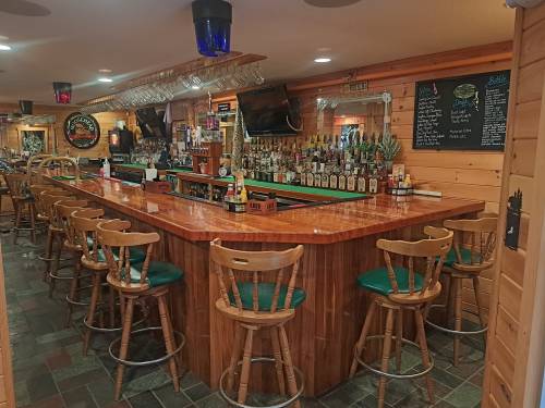 A wooden bar with high stools, lined with bottles, in a cozy, rustic setting with warm lighting.