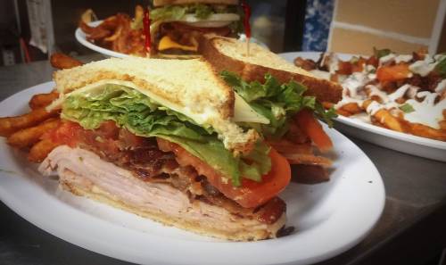A close-up of a sandwich with turkey, bacon, lettuce, and tomato, served with sweet potato fries in the background.