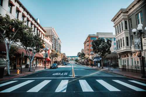 A quiet urban street with shops and trees, featuring a clear blue sky and a visible stop sign on the crosswalk.