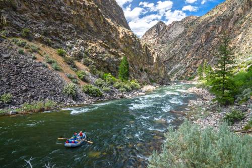 A group rafting on a river surrounded by steep, rocky mountains and lush greenery under a blue sky with clouds.