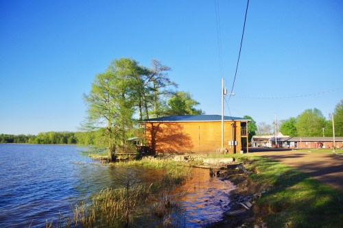 A wooden building by a calm lake, surrounded by greenery and a clear blue sky. Power lines run overhead.