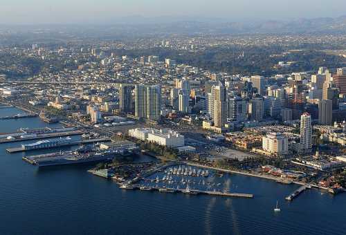 Aerial view of a coastal city with skyscrapers, marinas, and a waterfront, surrounded by hills in the background.