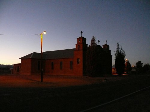 A silhouette of a church at dusk, with two towers and trees, illuminated by a streetlight.