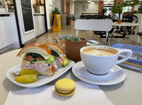 A sandwich with turkey, avocado, and tomato, a cup of latte, a macaron, and a small plant on a table in a café.