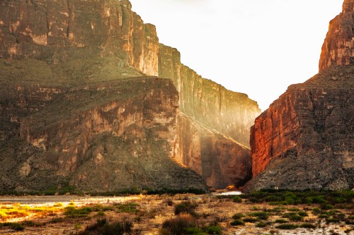Sunlight filters through steep canyon walls, illuminating a riverbed surrounded by rugged rock formations.