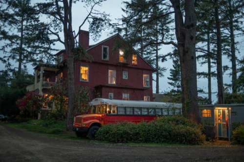 A red bus parked near a large, illuminated house surrounded by tall trees at dusk.