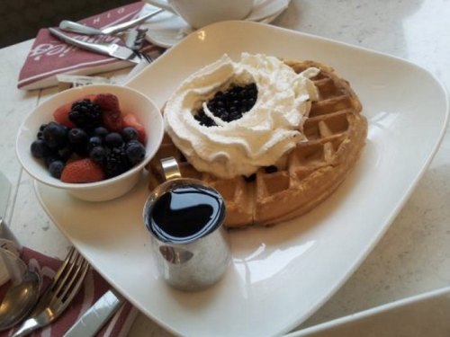 A plate with a waffle topped with whipped cream and berries, alongside a bowl of mixed berries and a small syrup container.