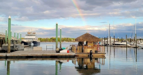 A serene marina scene with a thatched-roof hut, boats, and a rainbow in the sky reflecting on the calm water.