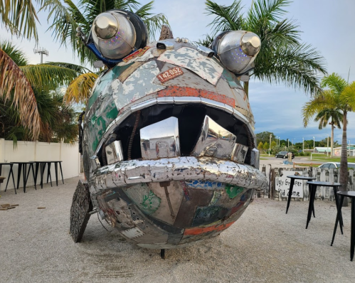 A large, whimsical fish sculpture made from metal scraps, featuring headlights for eyes and a wide, open mouth.