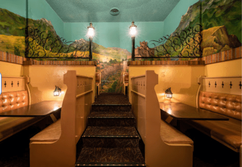 Cozy restaurant interior with booths, decorative murals, and stairs leading to an upper level. Warm lighting enhances ambiance.